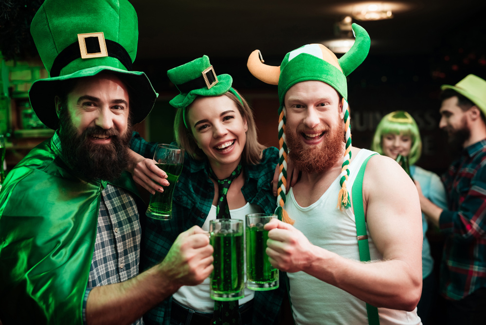 Two men and girl in carnival hats drinking beer at the bar. They celebrate St. Patrick's Day. They are having fun