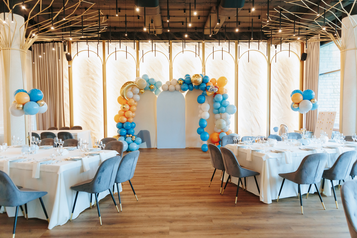 A beautifully decorated event venue featuring a balloon arch in blue, orange, and gold hues, elegant table settings with white tablecloths, and stylish modern chairs
