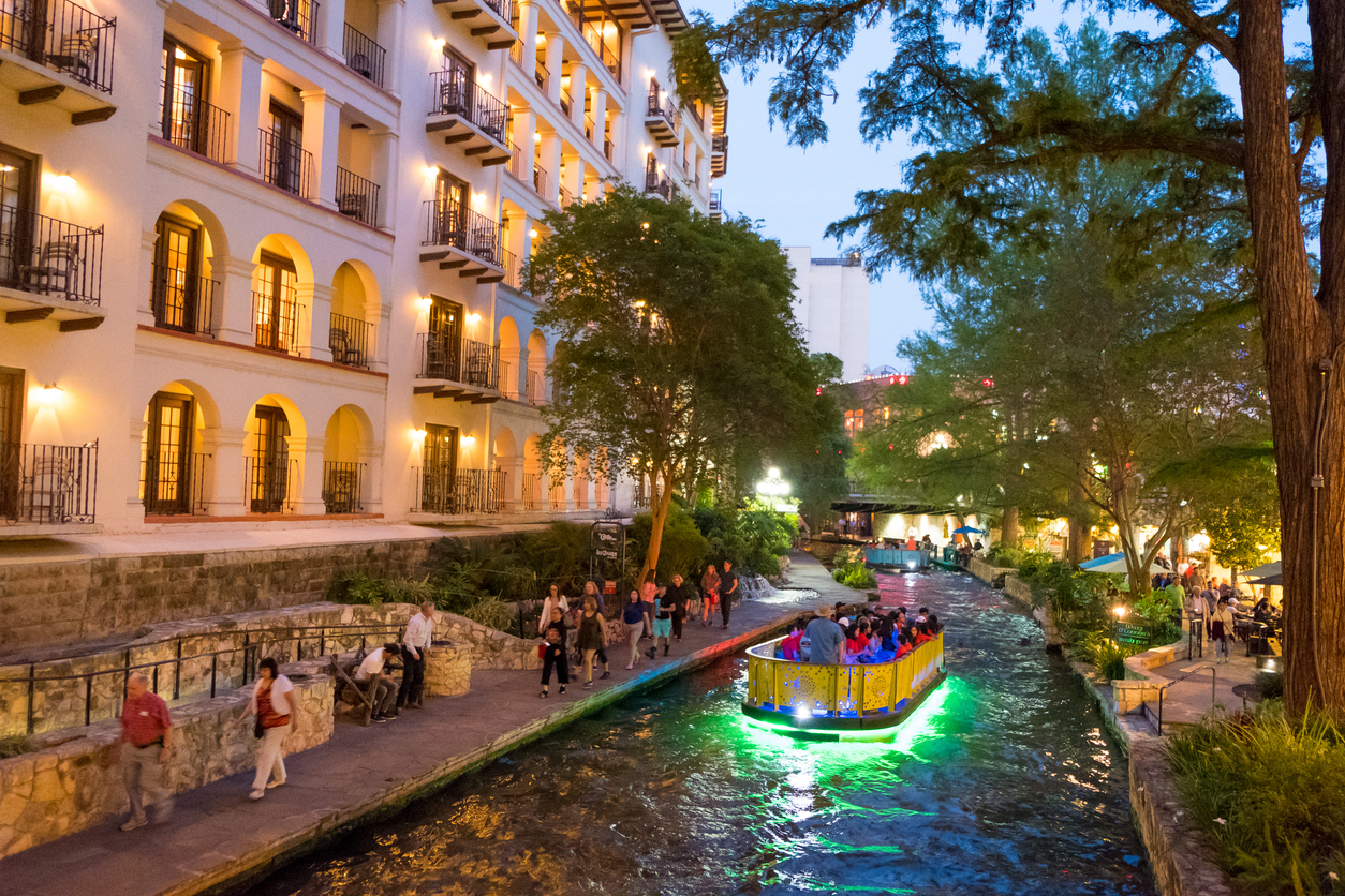 Boats passing through the river with pedestrians on the walkways of the River Walk in downtown San Antonio at twilight