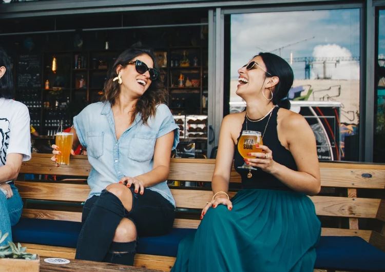 two laughing women sitting on a bench while holding a glass of beer