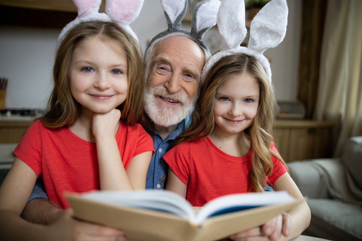 Cute twin girls with their happy grandfather sitting with bunny ears on heads and reading