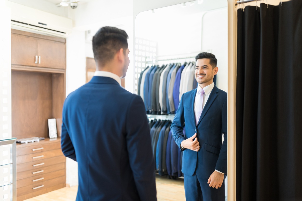 Reflection of Hispanic customer smiling while wearing new suit in rental store