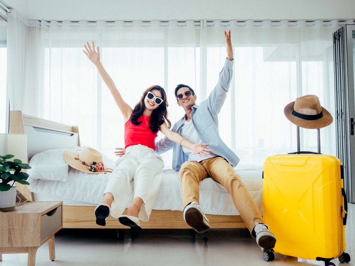 couple raising hands with joy, man and woman wear sunglasses smile with happy on white bed with yellow suitcases on a trip