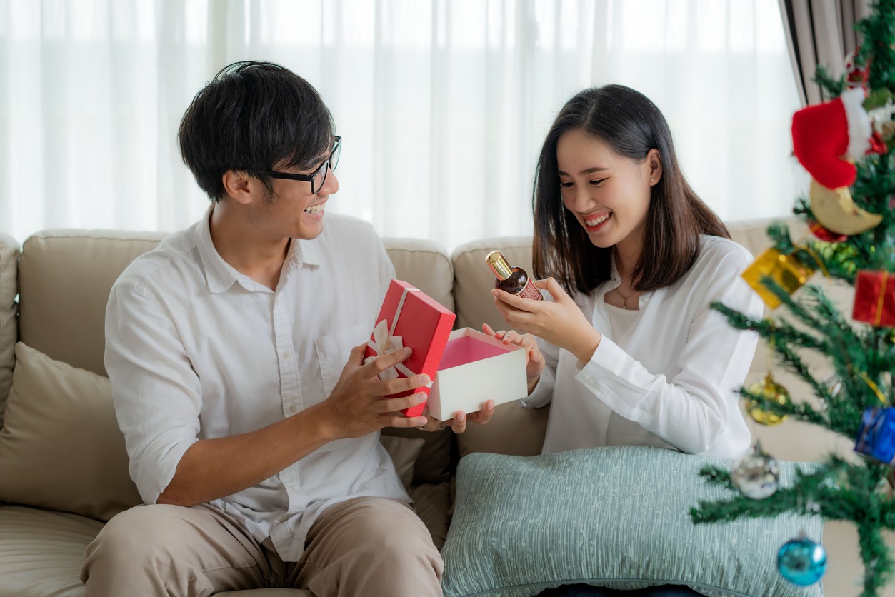 Asian man give the woman a red gift box in which there is a bottle of perfume. The woman was picked up on the sofa at home. The Christmas tree into the foreground. Love, relationship, wedding, or relaxing casual lifestyle concept