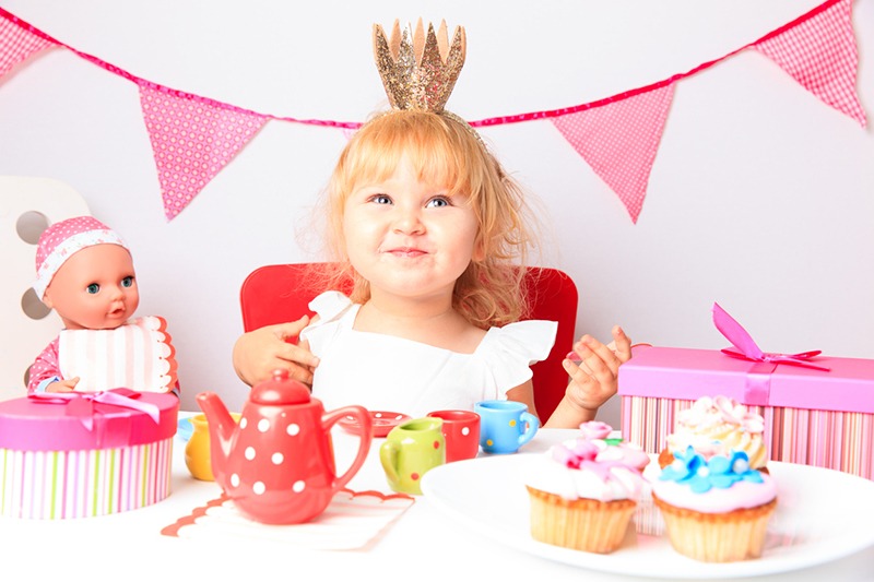 a smiling little girl wearing a crown in front of a table with sweets and dolls