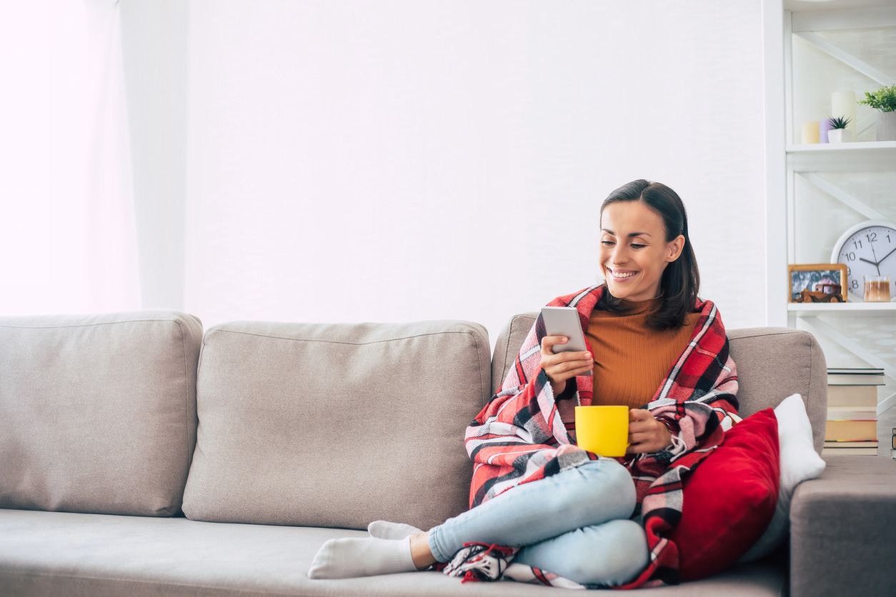 Happy young woman with a smartphone and cup in hands is covered under a checkered plaid on the couch while resting at home