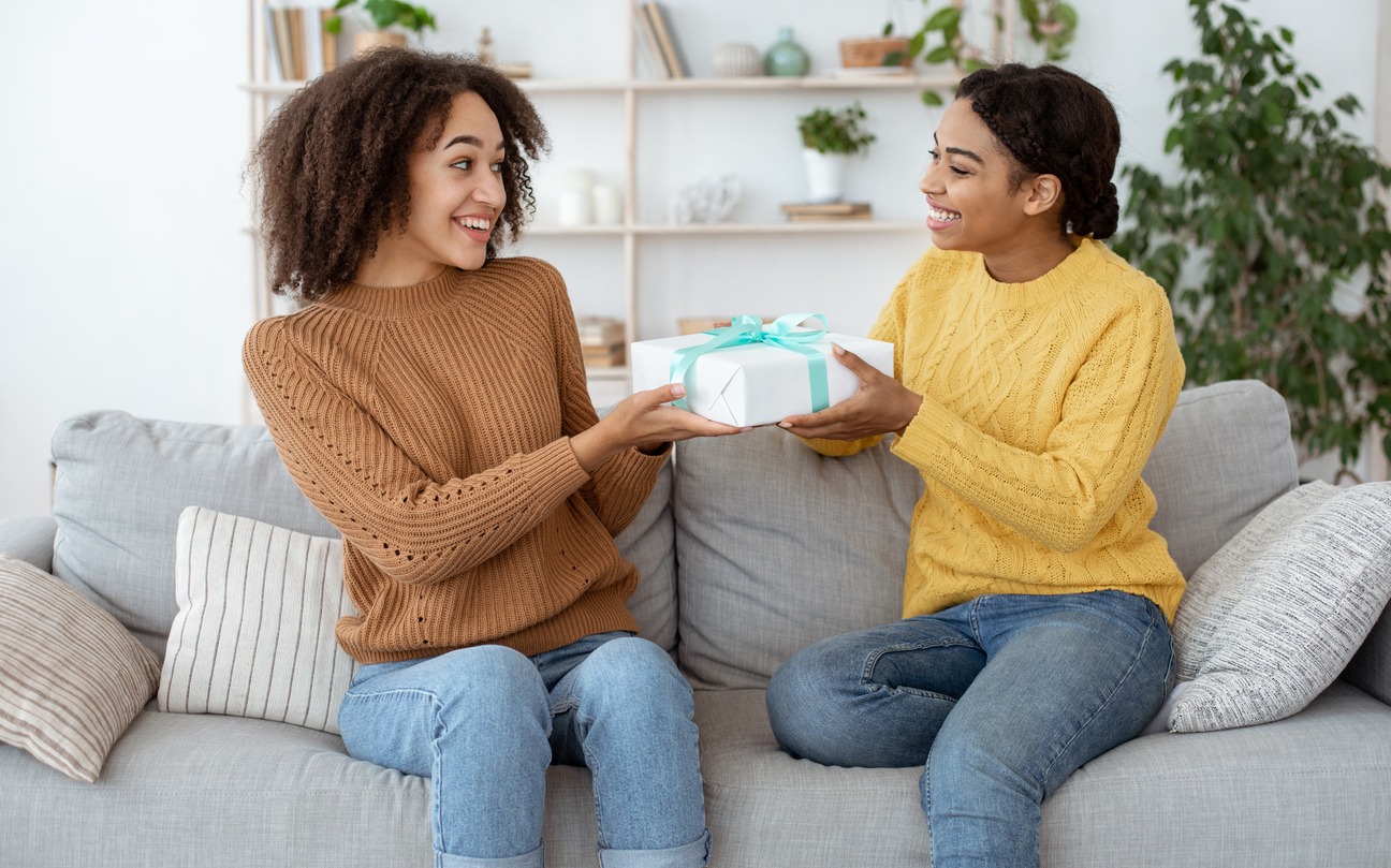 : Two ladies friends making surprise birthday present. Happy millennial african american female congratulates, gives gift to embarrassed mixed race sister, sits on sofa in interior of living room