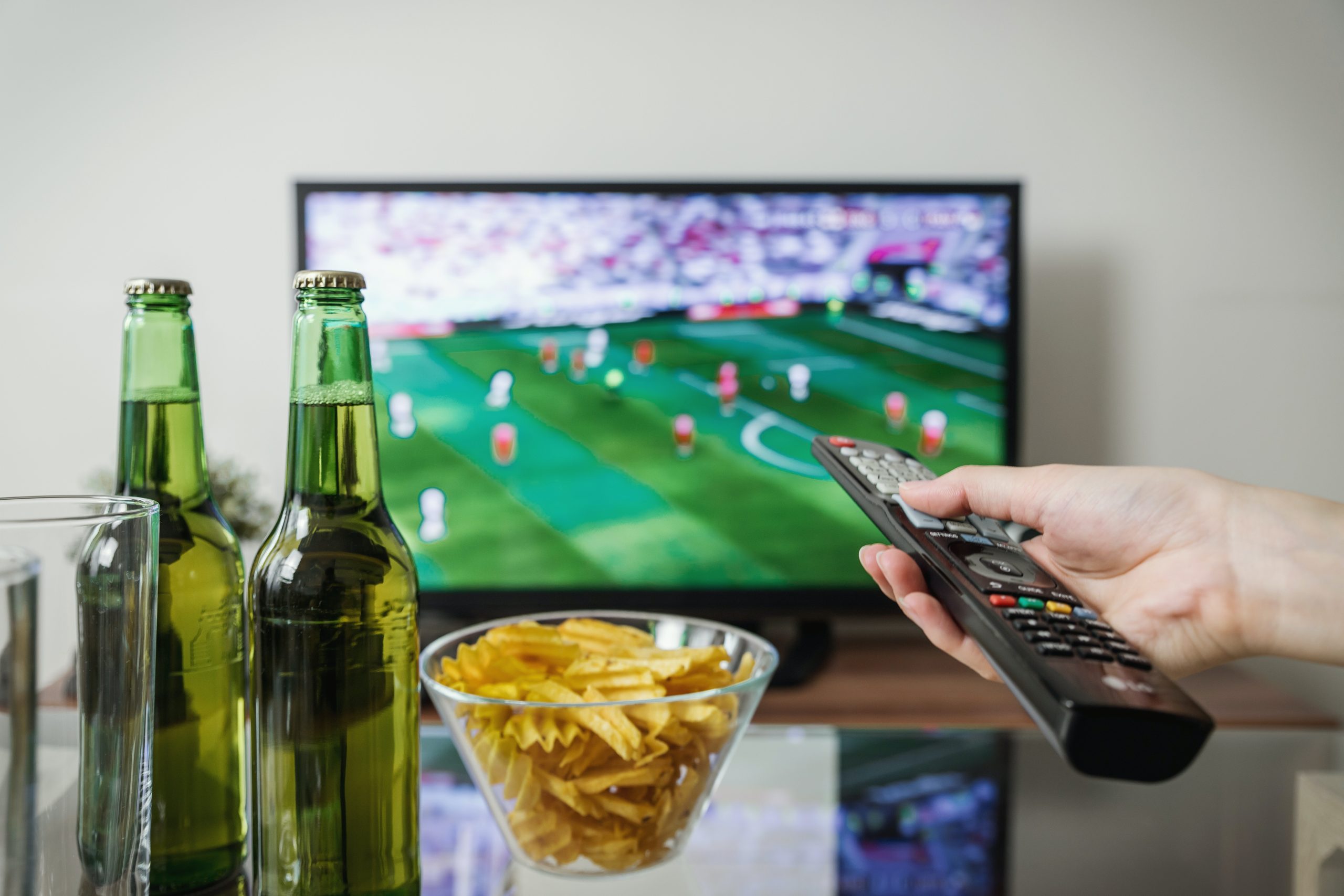 Hand holding the remote for the flat screen TV with chips and beer on the table 