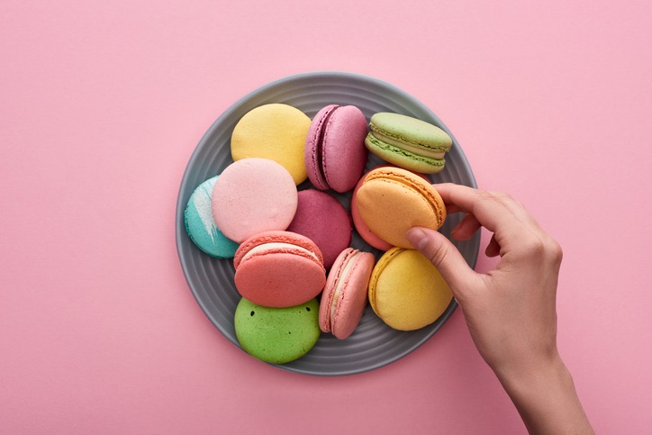 cropped view of female hand near plate with multicolored delicious French macaroons on pink background