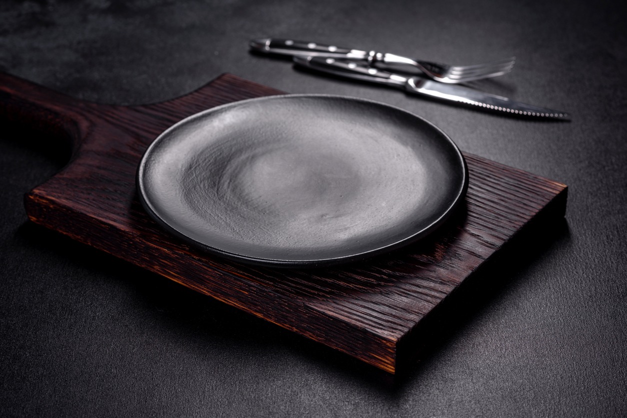  An empty plate with a knife, fork or spoon with a wooden cutting board on a dark concrete background. Preparation of appliances and ingredients for home cooking