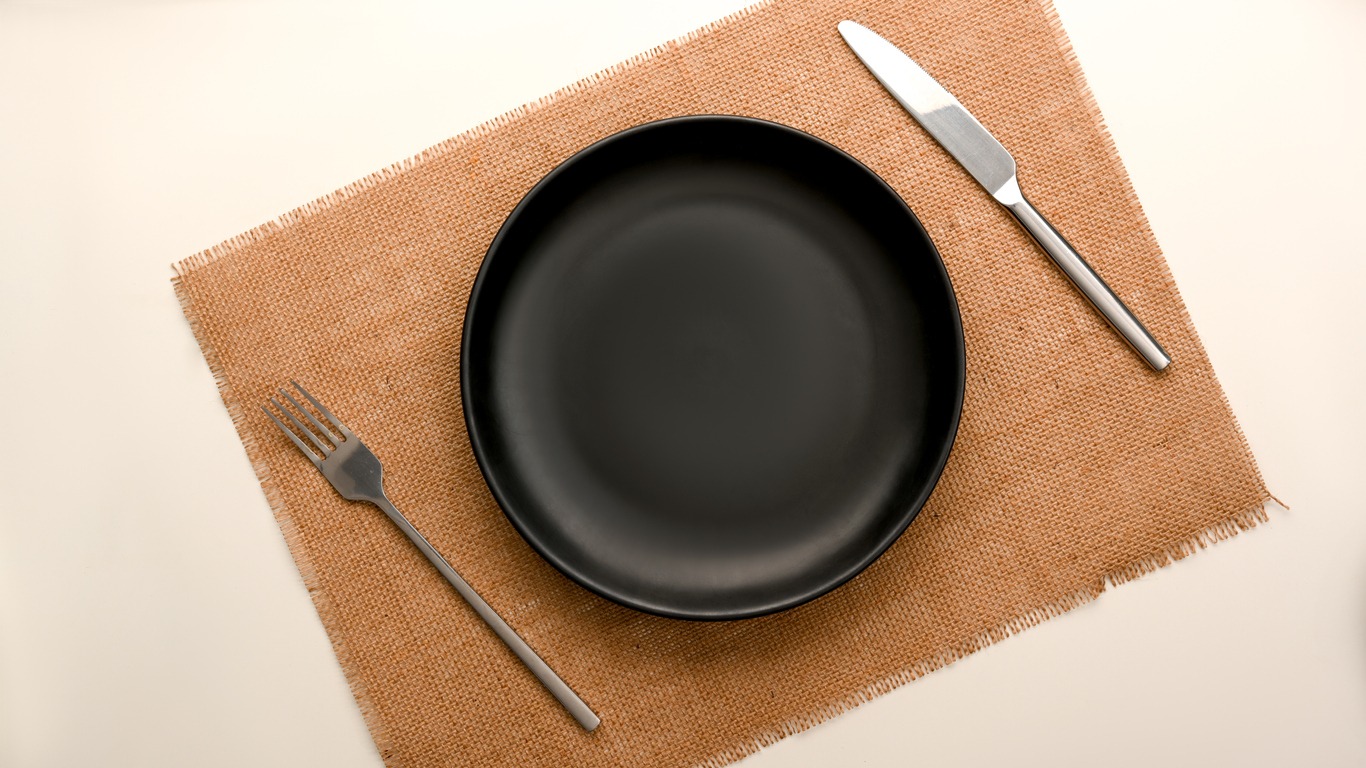 Top view, flat lay, a minimal dining table setting with empty black plate mockup and silverware over placemat.