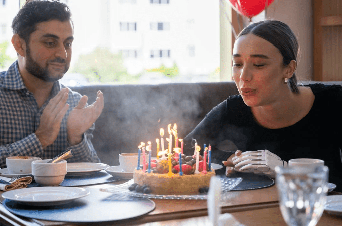 a woman sitting beside a clapping man in a restaurant blowing her birthday candles