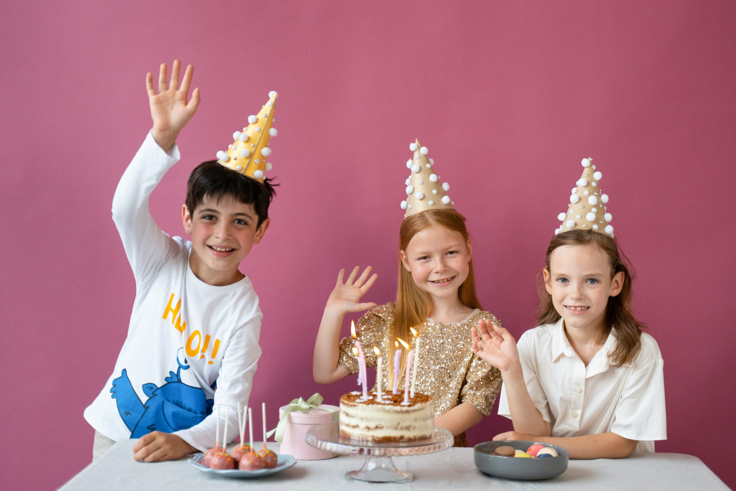 three children wearing party hats in front of a table with a cake and other desserts on top