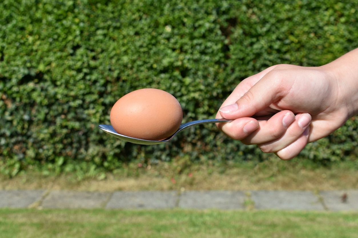Close up of hard-boiled egg in shell on metal spoon held by a woman's hand outside in summer in front of a green hedge- being used for egg and spoon race