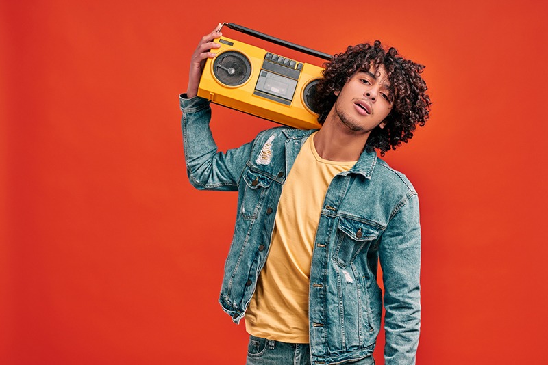 Waist up portrait of young handsome stylish man with bright beat box on his shoulder on isolated orange