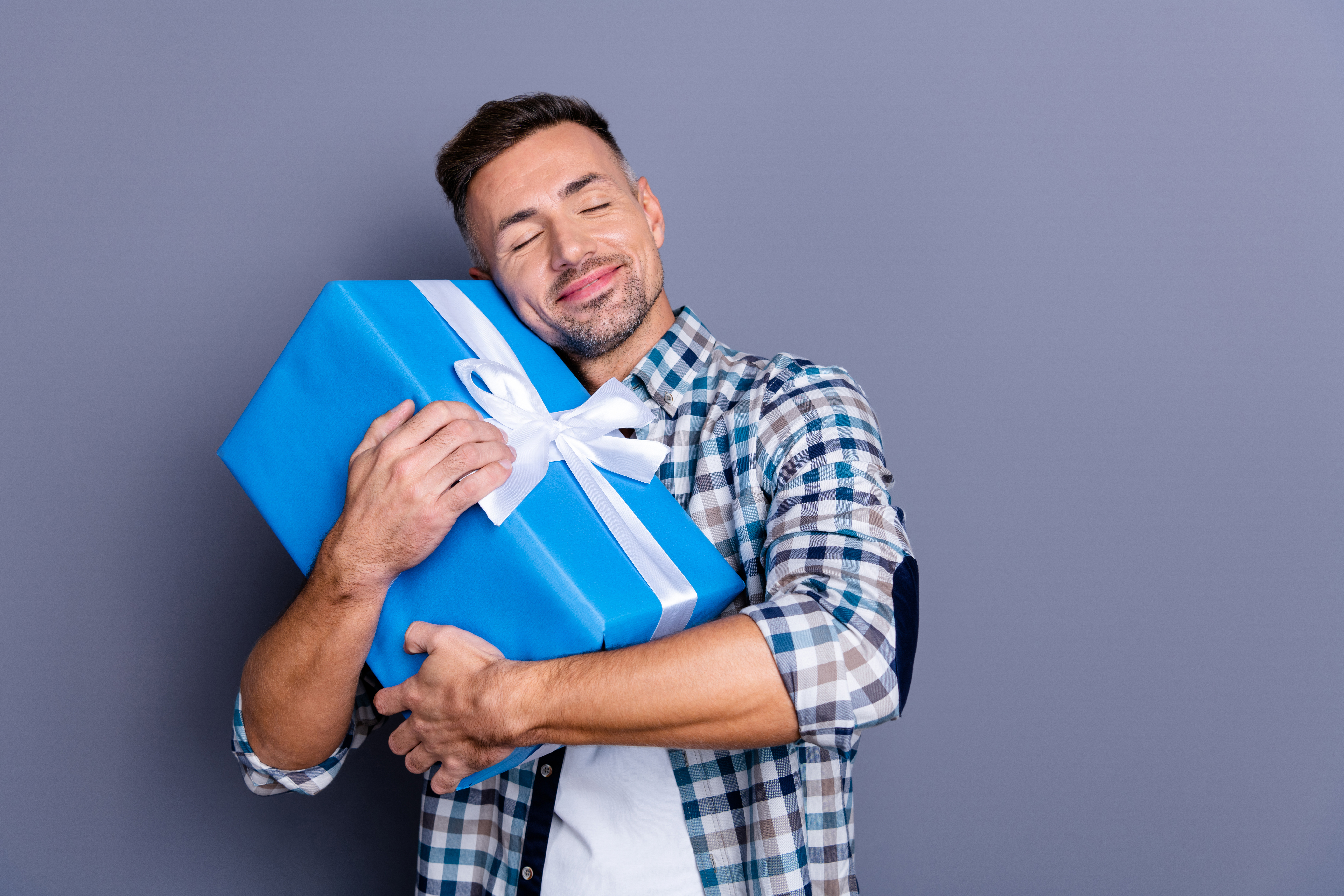 Portrait of nice cheerful bearded guy wearing checked, shirt holding in hands hugging festive celebratory box isolated over blue violet purple pastel background