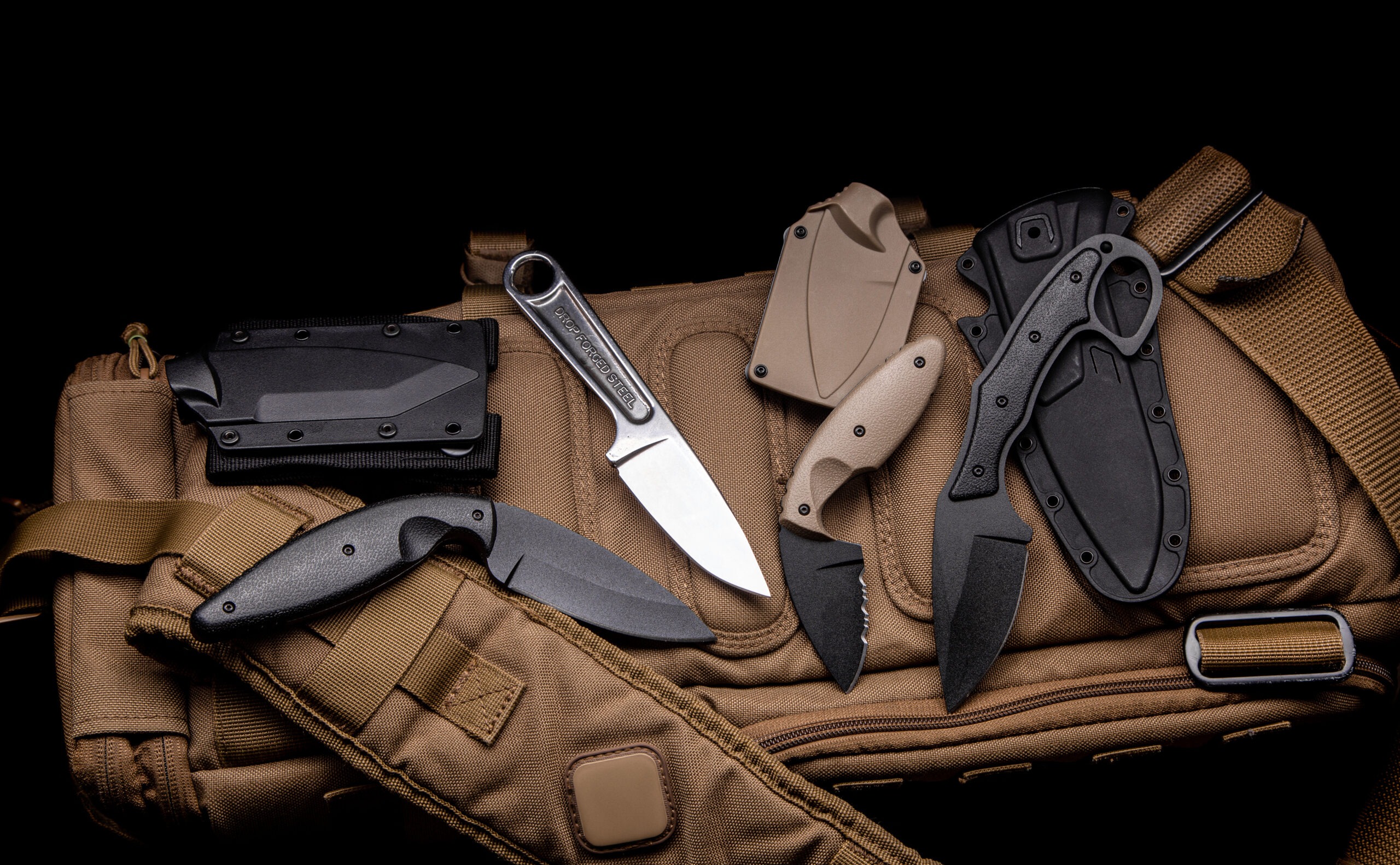 A variety of knives on a sand-colored military backpack. Dark background